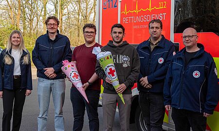 Eine junge Frau mit langen, hellen Haaren, die ein dunkles Jackett trägt. Daneben stehen fünf Männer, von denen zwei in Uniformen des DRK gekleidet sind. Die beiden Personen in der Mitte halten jeweils einen bunt gestalteten Konus, der typischerweise bei Geburtstagen oder Feiern im DRK-Bereich verwendet wird. Die Konusse tragen die Namen „DANI“ und „ELIAS“ sowie das Logo des DRK. Die Gruppe wirkt freundlich und posiert gemeinsam für das Foto. Im Hintergrund sind Bäume und der Rettungswagen zu sehen.