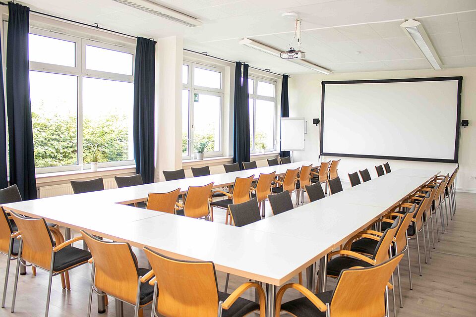 Ein großer Seminarraum mit rechteckiger Tischanordnung, vielen Stühlen, Projektionsleinwand und Tageslicht durch mehrere Fenster.