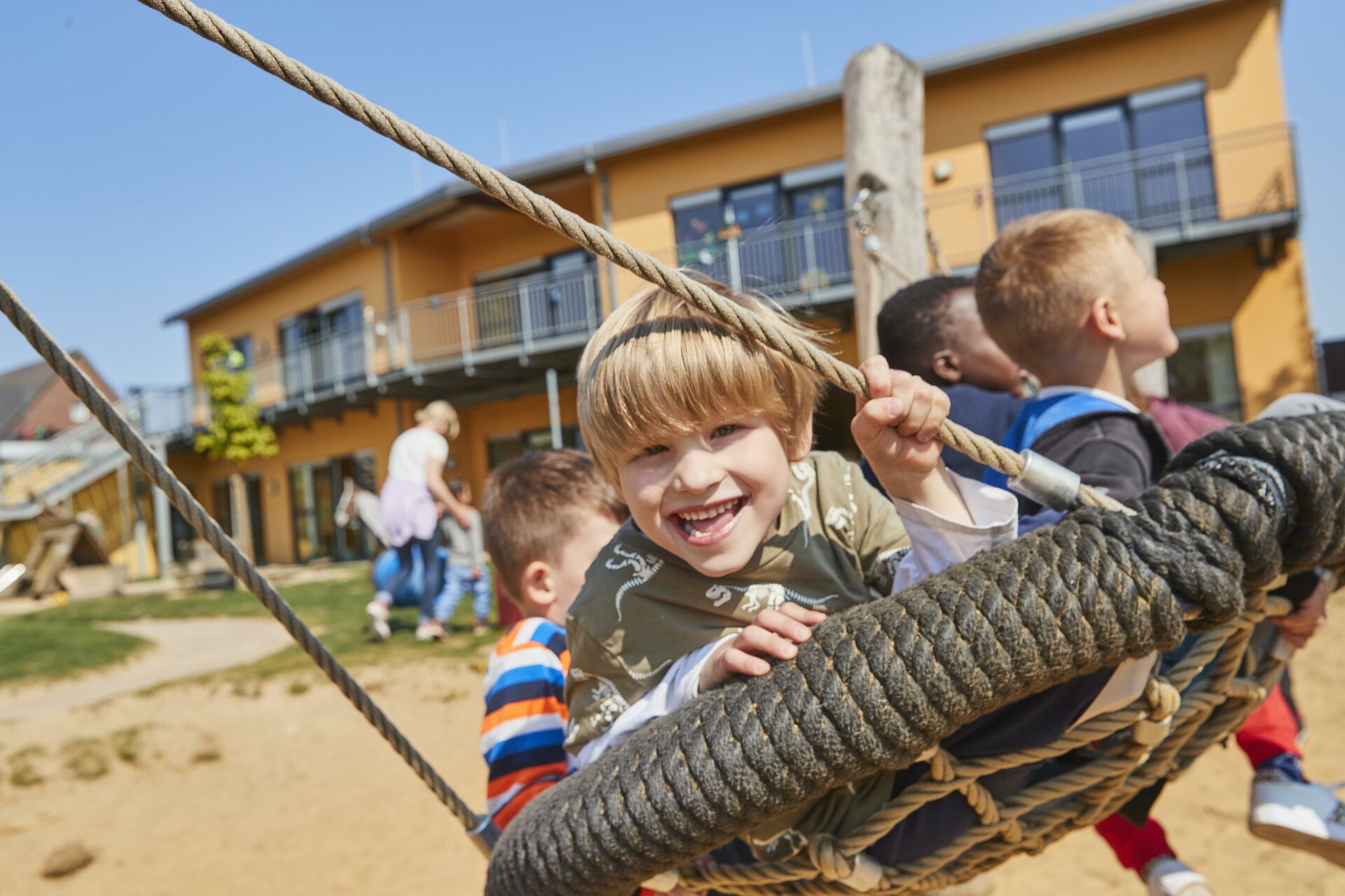 Ein lachender Junge mit blonden Haaren sitzt auf einer Seilschaukel und hält sich an einem Seil fest, während im Hintergrund weitere Kinder spielen und ein orangefarbenes Gebäude zu sehen ist.