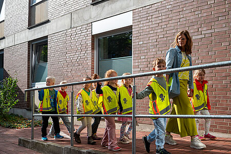 Eine Gruppe von Kindergartenkindern geht mit einer Erzieherin an einem Backsteingebäude entlang. Alle Kinder tragen leuchtend gelbe Sicherheitswesten mit reflektierenden Streifen und Symbolen. Die Erzieherin läuft vorne rechts, hält ein Kind an der Hand und trägt eine gelbe Hose mit einer Jeansjacke. Die Kinder gehen in einer Reihe, einige schauen zur Seite, andere nach vorne.