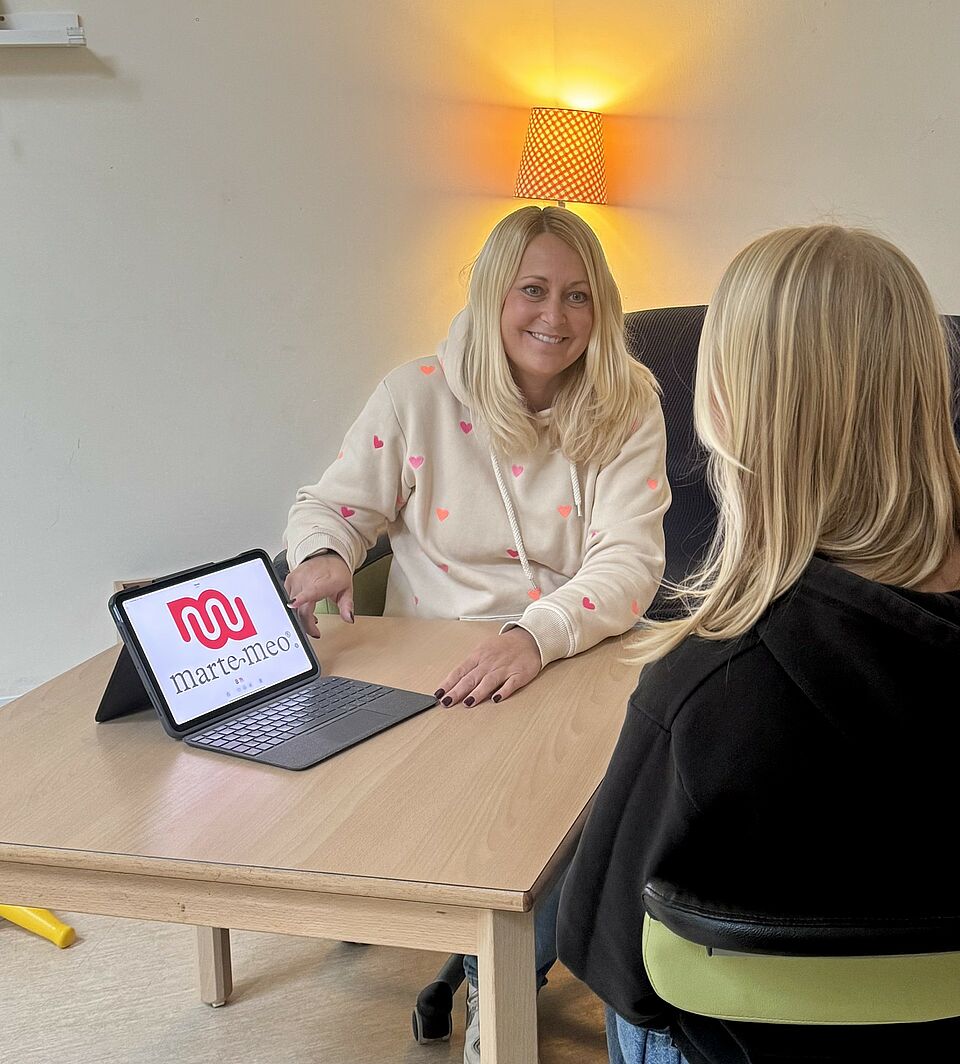 Zwei Frauen sitzen gemeinsam an einem Holztisch in einem hellen, gelb gestrichenen Raum mit weißer Wand. Die Frau links trägt ein helles, kariertes Hemd mit roten und pinken Akzenten und hält ein Tablet mit einem Bildschirm, auf dem teilweise die Website „haut.maremeo.“ sichtbar ist. Sie hat blonde, leicht gewellte Haare und lächelt freundlich. Die Frau rechts trägt eine dunkle Brille und einen dunklen, glatten Pullover. Im Hintergrund ist ein weiterer Bildschirm an der Wand montiert, daneben ein Regal oder Schrank mit Gegenständen. Auf dem Tisch liegen ein Handy und ein Kabel.