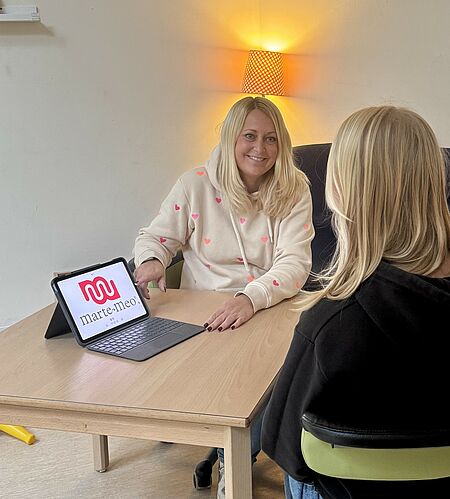 Zwei Frauen sitzen gemeinsam an einem Holztisch in einem hellen, gelb gestrichenen Raum mit weißer Wand. Die Frau links trägt ein helles, kariertes Hemd mit roten und pinken Akzenten und hält ein Tablet mit einem Bildschirm, auf dem teilweise die Website „haut.maremeo.“ sichtbar ist. Sie hat blonde, leicht gewellte Haare und lächelt freundlich. Die Frau rechts trägt eine dunkle Brille und einen dunklen, glatten Pullover. Im Hintergrund ist ein weiterer Bildschirm an der Wand montiert, daneben ein Regal oder Schrank mit Gegenständen. Auf dem Tisch liegen ein Handy und ein Kabel.