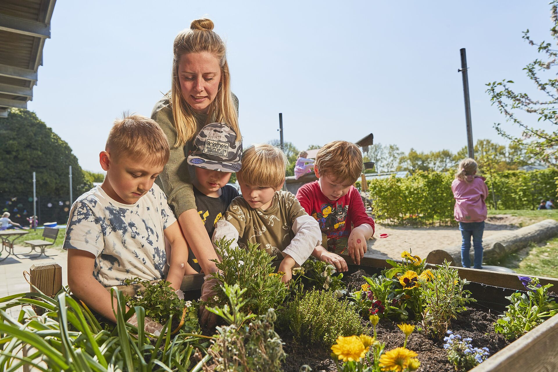 Eine Frau und vier Kinder pflanzen gemeinsam Blumen und Kräuter in einem Hochbeet im Freien.