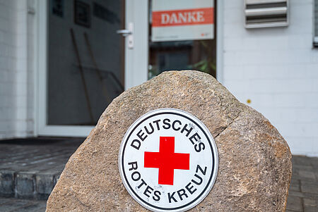 Ein Stein mit dem runden Logo des Deutschen Roten Kreuzes steht im Vordergrund. Im Hintergrund ist der Eingang eines DRK-Gebäudes zu sehen, mit einer Glastür und einem Schild mit der Aufschrift „Danke“.