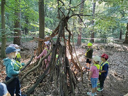 Waldtage Wiesenzwerge Kinder der DRK-Kita 'Wiesenzwerge' aus Ibbenbüren bauen gemeinsam ein Tipi aus Ästen im Wald. Sie tragen Warnwesten und Mützen und sind von Bäumen und Laub umgeben.