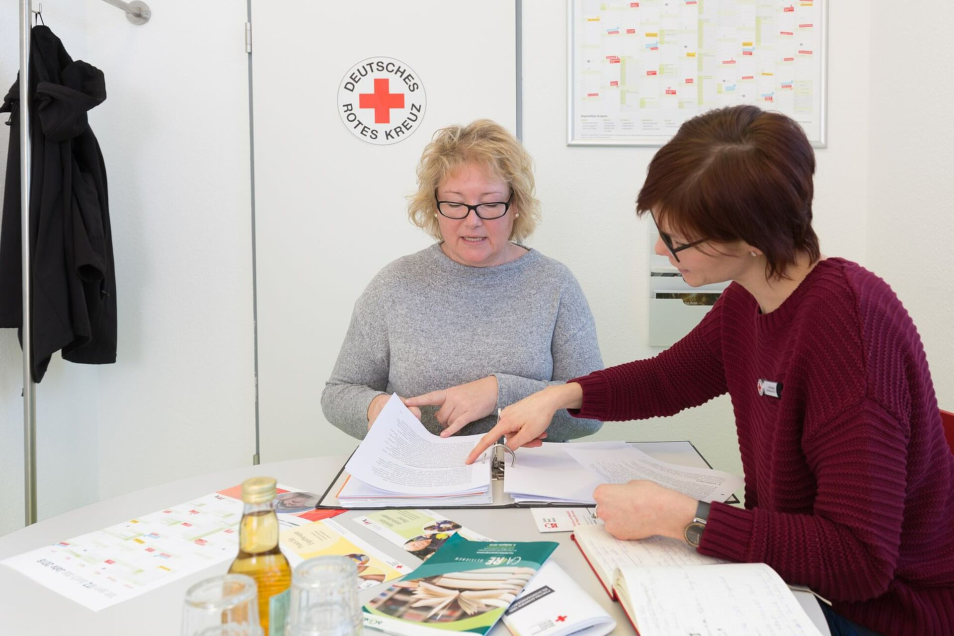 Zwei Frauen sitzen an einem Schreibtisch und besprechen Unterlagen, wobei eine Frau auf ein Dokument zeigt. Im Hintergrund ist das Logo des Deutschen Roten Kreuzes zu sehen.