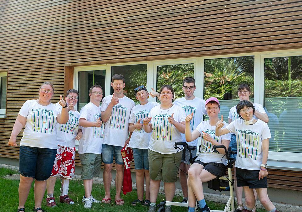 Gruppe von neun Personen, die vor einem Holzgebäude mit großen Fenstern stehen. Alle tragen weiße T-Shirts mit der Aufschrift '10 Jahre Lachgesichter' und bunte Muster. Einige zeigen den Daumen nach oben, andere lächeln in die Kamera. Eine Person sitzt in einem Rollstuhl, eine weitere trägt eine rote Basecap. Im Hintergrund sind grüne Sträucher und Bäume zu sehen.