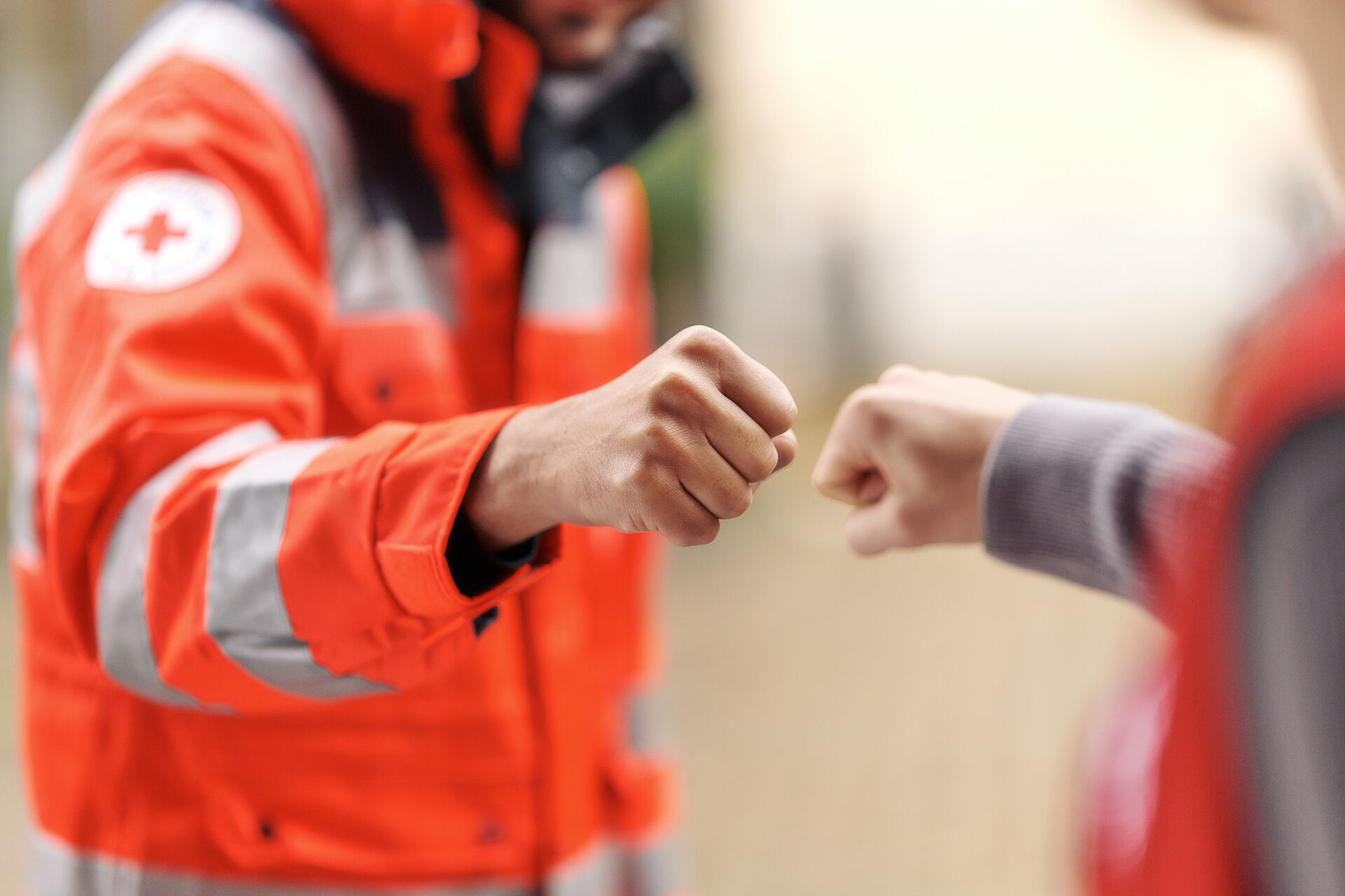 Zwei Personen machen einen Fauststoß, eine davon trägt eine orangefarbene DRK-Einsatzjacke.