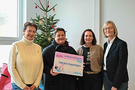 Vier Frauen stehen in einem hellen Raum vor einem geschmückten Weihnachtsbaum. Die zweite Frau von links hält ein Schild mit der Aufschrift „Zertifizierter Kindergarten – DRK-KiTa Regenbogen“. Alle lächeln in die Kamera. Die Frauen tragen winterliche, dezente Kleidung, und die Szene wirkt freundlich und feierlich.