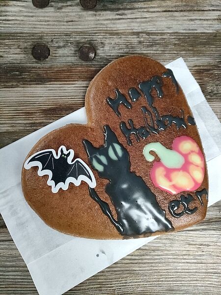 Ein Lebkuchenherz liegt auf einer Serviette auf einem Holztisch. Es ist mit schwarzem und buntem Zuckerguss zu Halloween-Motiven verziert – darunter eine schwarze Katze, eine Fledermaus, ein Kürbis und der Schriftzug „Happy Halloween“.