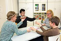Vier Personen, darunter zwei Jugendliche und zwei Erwachsene, sitzen an einem Tisch und interagieren miteinander, wobei sie ihre Hände in spielerischer oder kommunikativer Geste einsetzen.