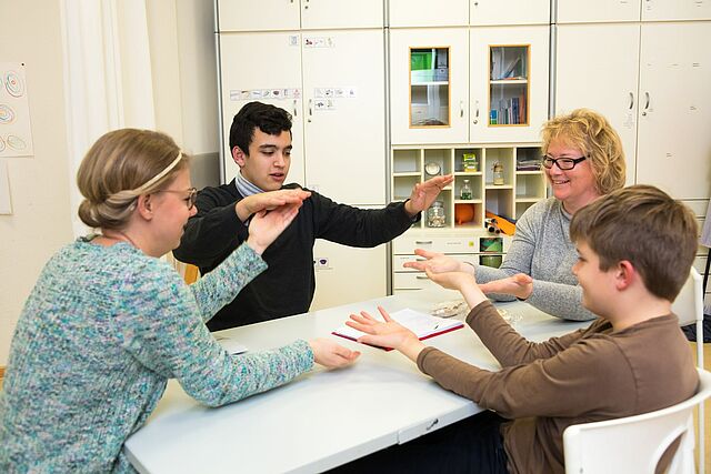 Vier Personen, darunter zwei Jugendliche und zwei Erwachsene, sitzen an einem Tisch und interagieren miteinander, wobei sie ihre Hände in spielerischer oder kommunikativer Geste einsetzen.