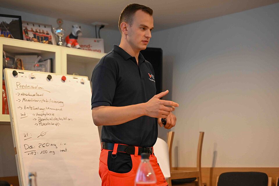 Ein Mann in dunkelblauem Poloshirt mit rotem Einsatzhosenteil steht vor einem Whiteboard und erklärt Inhalte. Auf dem Whiteboard sind handschriftliche Notizen zu Medikamenten zu sehen.